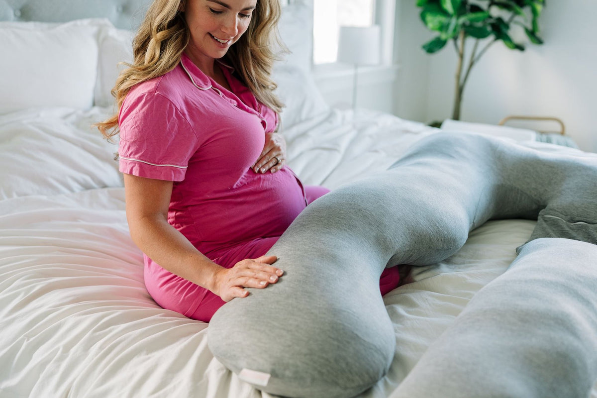 Pregnant woman in pink pajamas sitting on a bed with her hand on a gray Araluna U-shaped pregnancy pillow