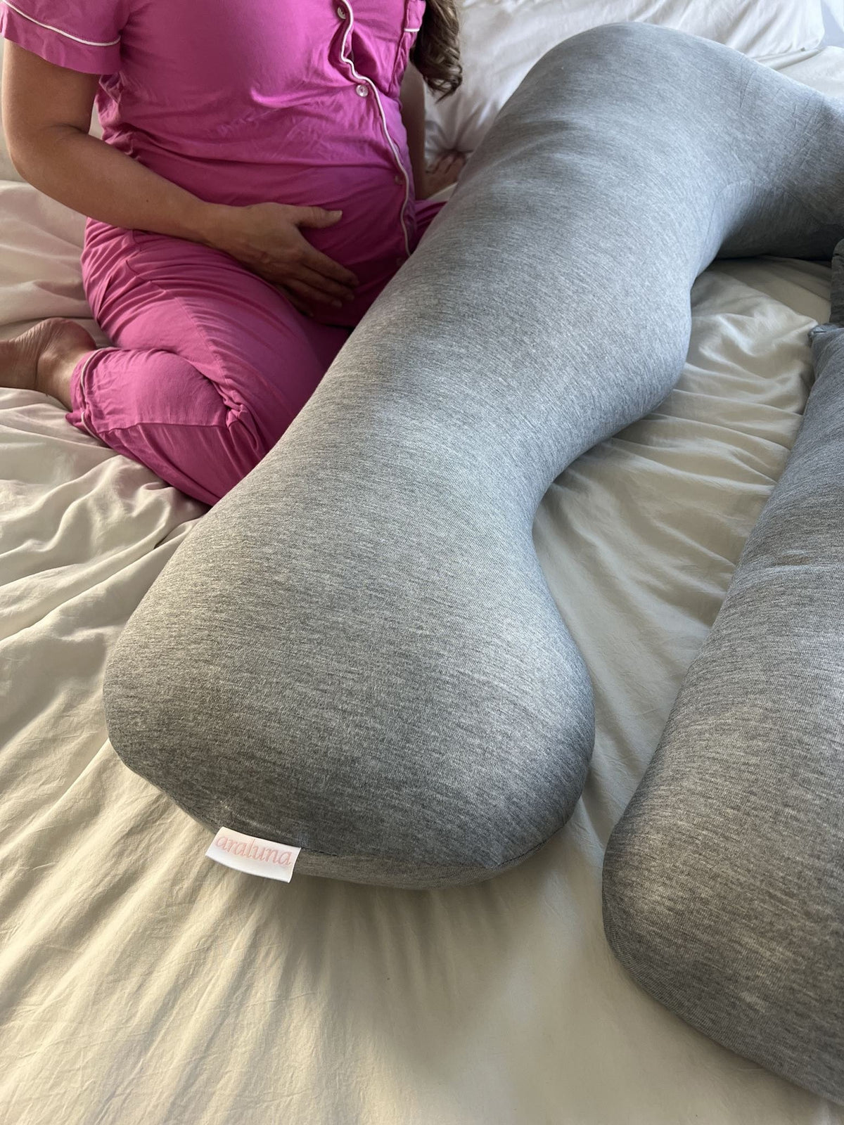 Pregnant woman sitting on a bed beside a gray Araluna pregnancy pillow